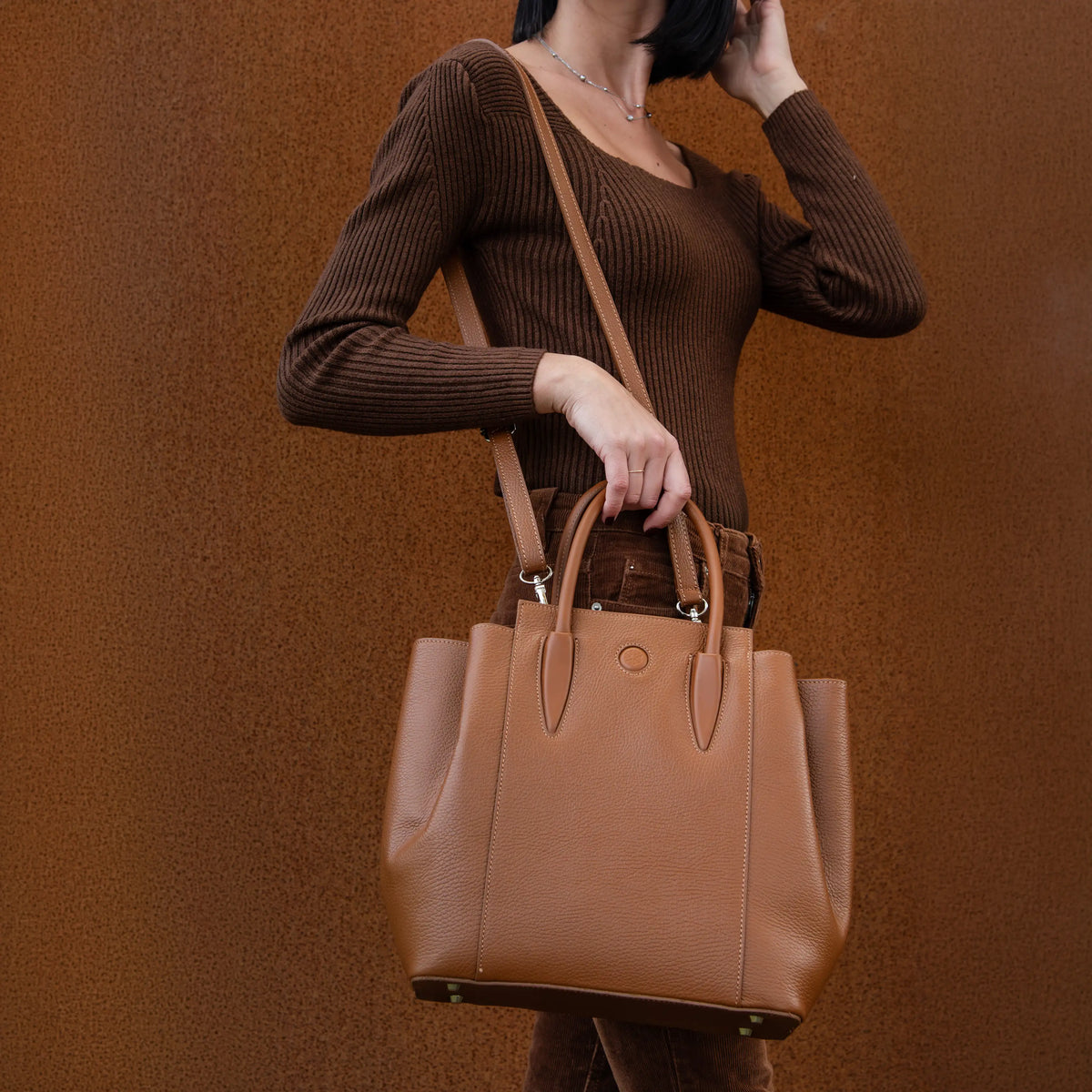 Person wearing a brown outfit carrying the Tulipan cognac brown handbag by the shoulder strap against a brown wall.