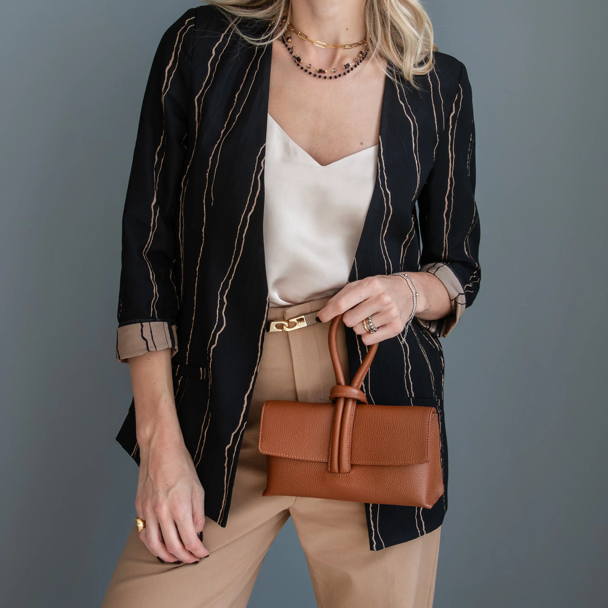A woman models a black three-quarter sleeve blazer with thin gold vertical stripes, paired with a cream camisole, tan trousers, and a cognac leather clutch handbag against a solid gray background.