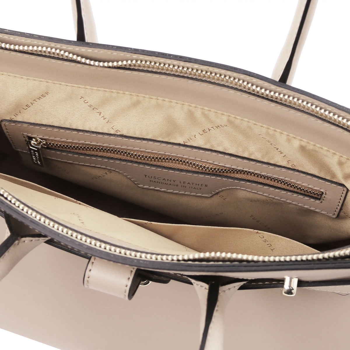 Close-up of the interior of a beige handbag, showing a zippered pocket with a leather label that reads 'TUSCANY LEATHER' and a cream-colored exterior edge with black trim.