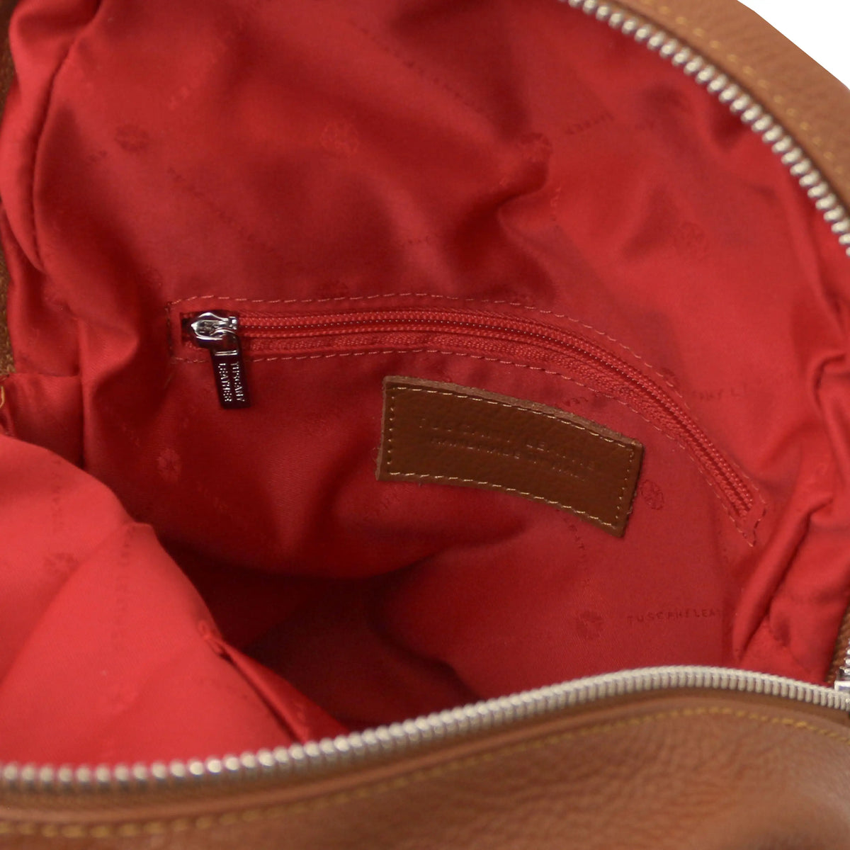 Close-up of the interior of a cognac backpack showing the red quilted lining, a zippered pocket with a silver zipper pull, and a brown leather brand patch.