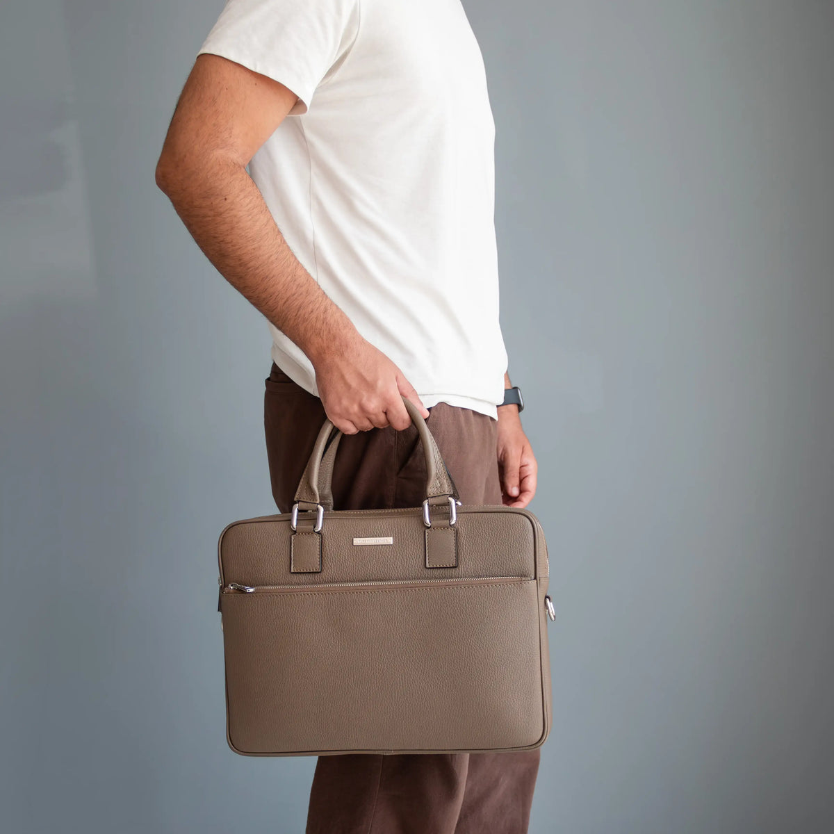 A man in a white t-shirt and brown trousers holds the Ferrara taupe pebbled leather briefcase with silver hardware against a solid gray background.