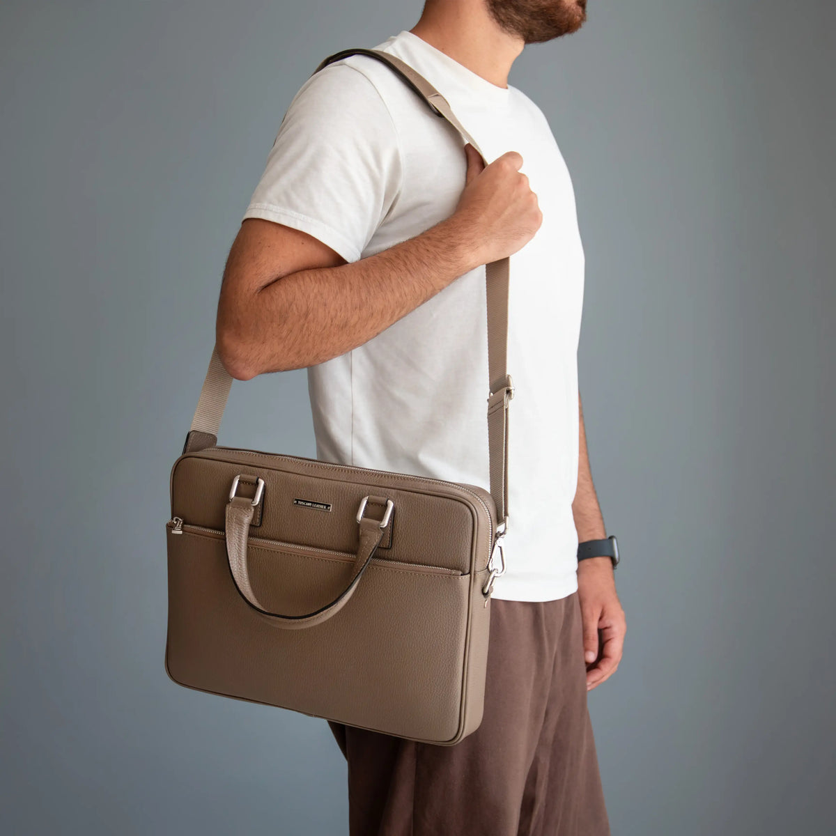 A man wearing a white t-shirt and brown trousers carries the Ferrara taupe leather briefcase-style laptop bag with a shoulder strap and top handle against a plain gray studio background