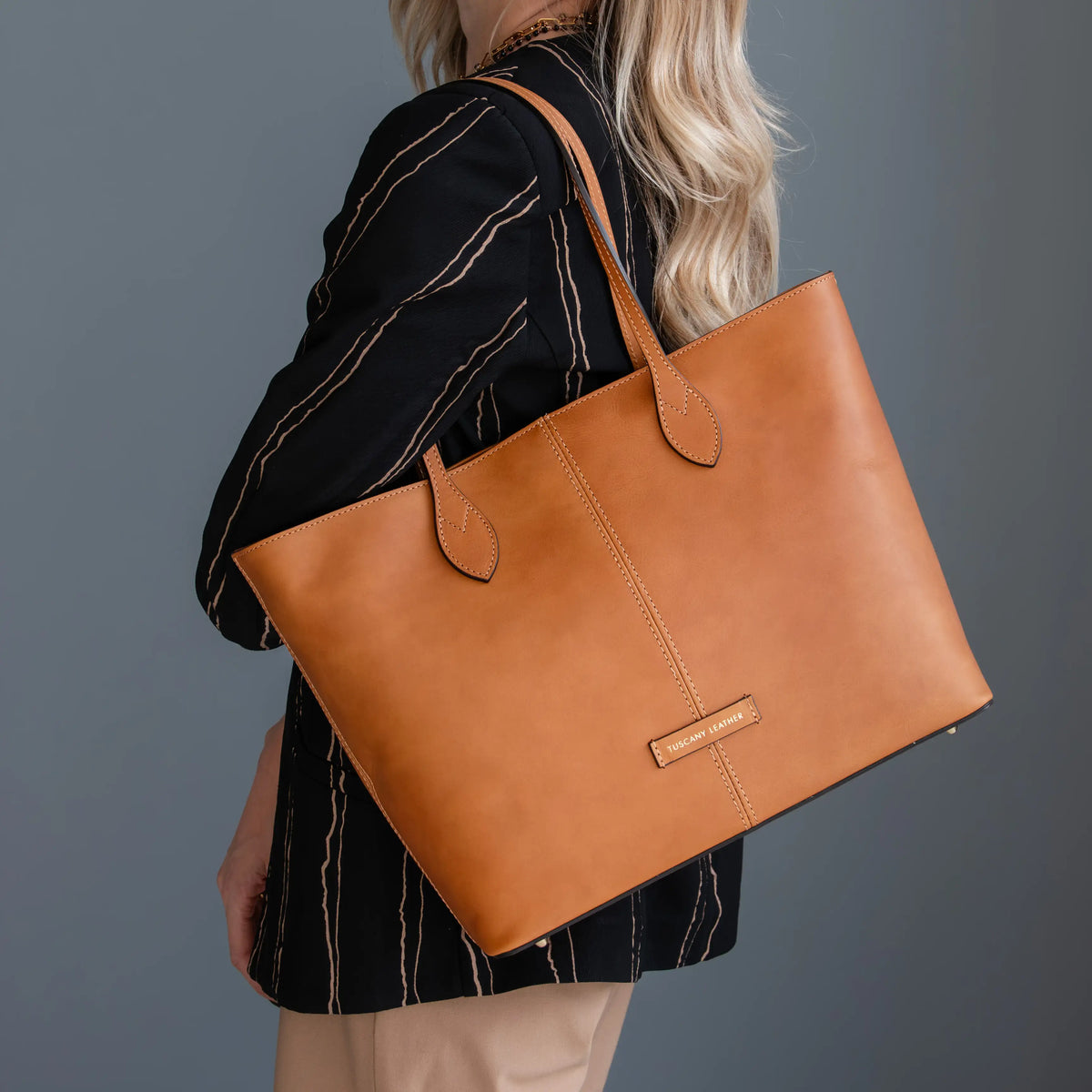 A woman wearing a black and beige striped blazer carries a large natural leather tote bag over her shoulder against a dark gray studio background.