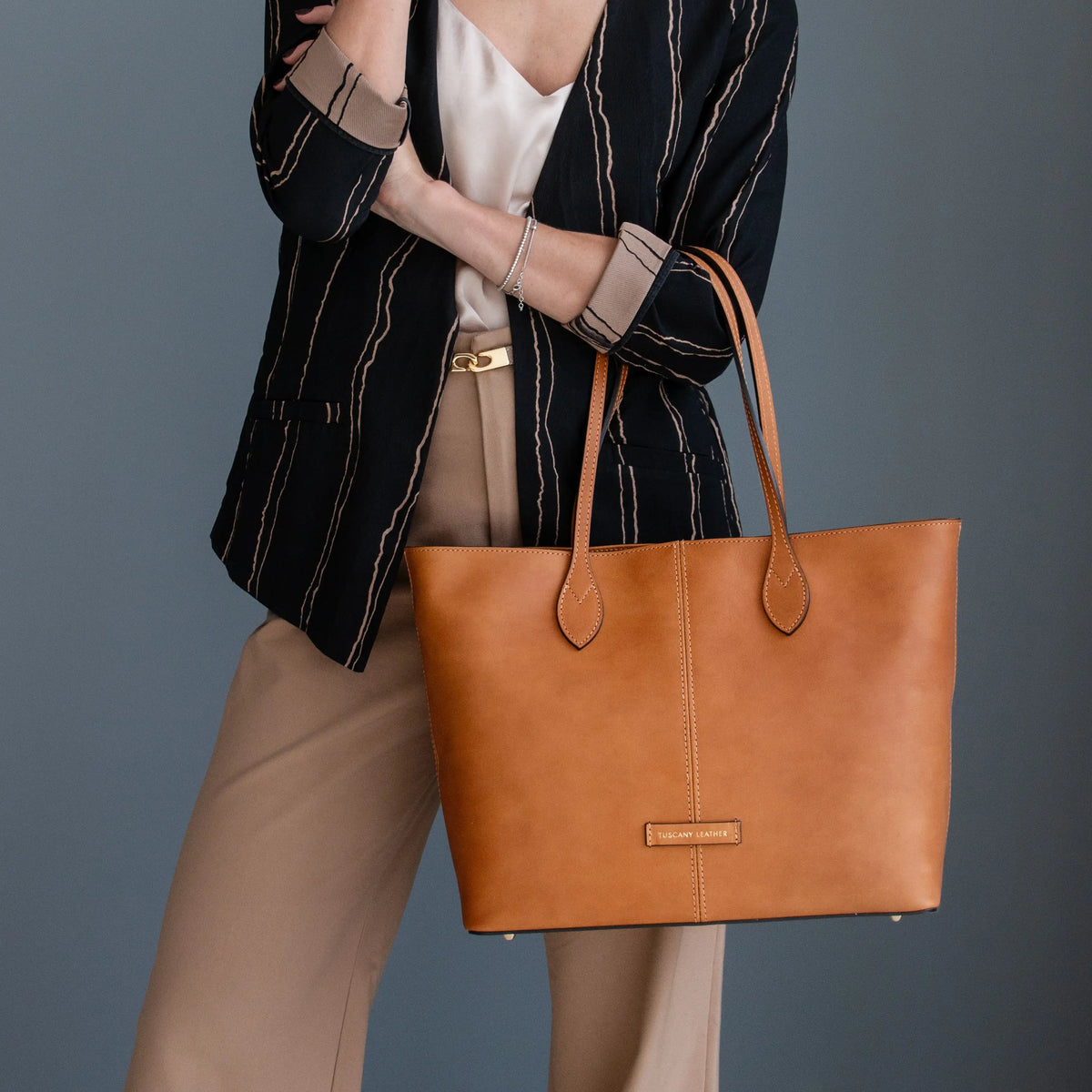 A woman wearing a black and beige striped blazer carries a large natural leather tote bag on her arm against a dark gray studio background.