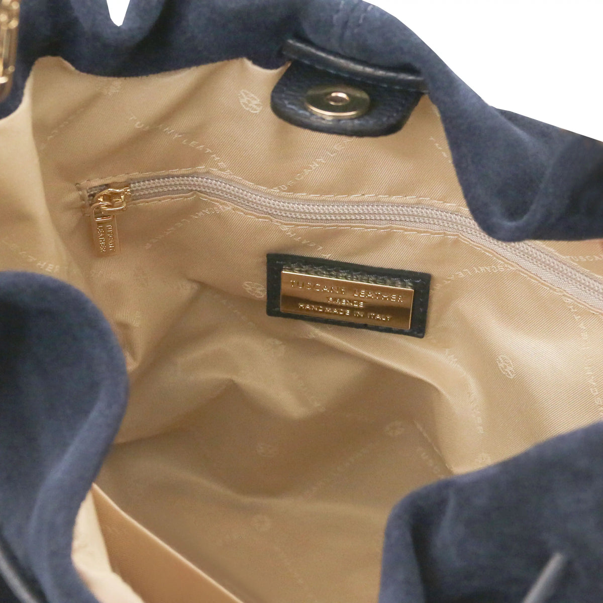 Close-up of the interior of a dark blue suede handbag, showing a beige quilted lining, a gold-tone zipper, and a black leather and gold metal brand label.
