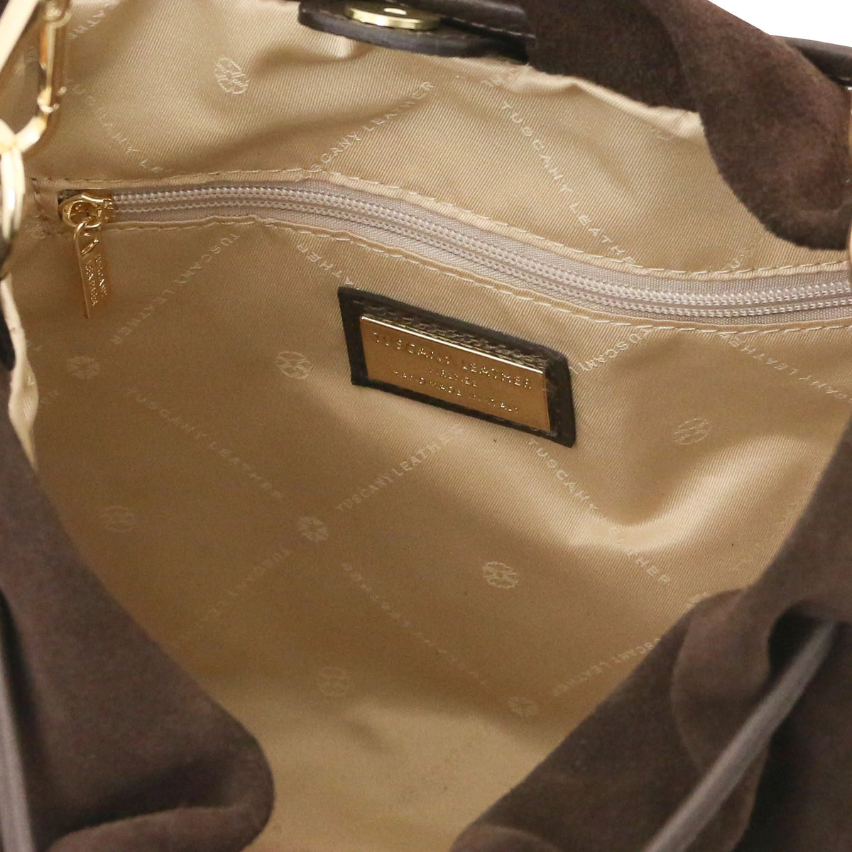 Close-up of the interior of a handbag showing a dark blue quilted nylon lining with a gold-tone logo plaque, a zippered pocket, and dark brown suede-like trim.