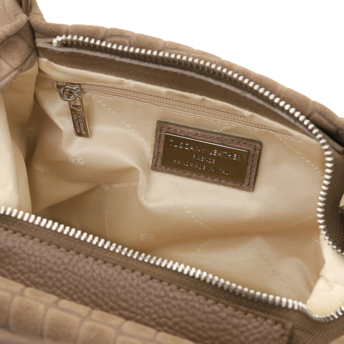 Close-up of the interior of a taupe croc textured handbag, showing a cream-colored lining, a silver zipper, and a brown leather brand patch.