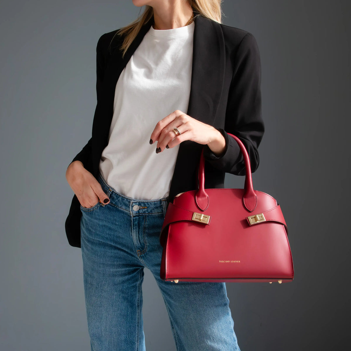 A woman wearing a black blazer, white t-shirt, and blue jeans holds a red leather handbag with gold hardware against a dark gray studio background.