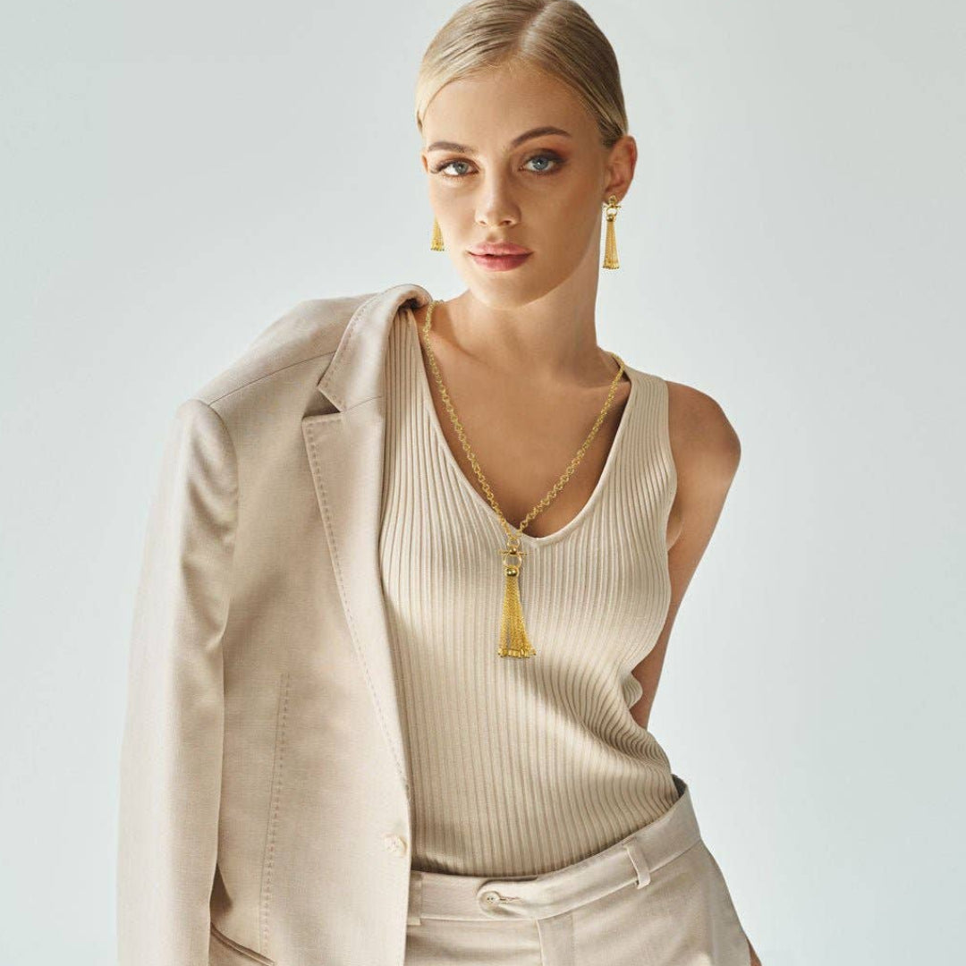 Woman wearing a beige outfit with a gold necklace and earrings on a light gray background