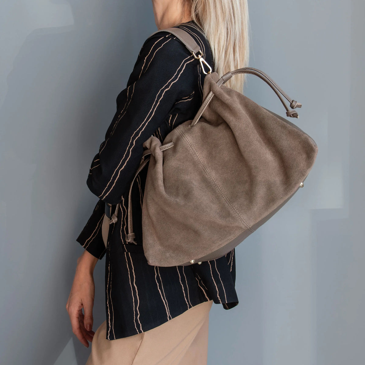 A woman wearing a black and beige striped shirt carries a large dark taupe suede drawstring shoulder bag against a muted blue-gray wall.