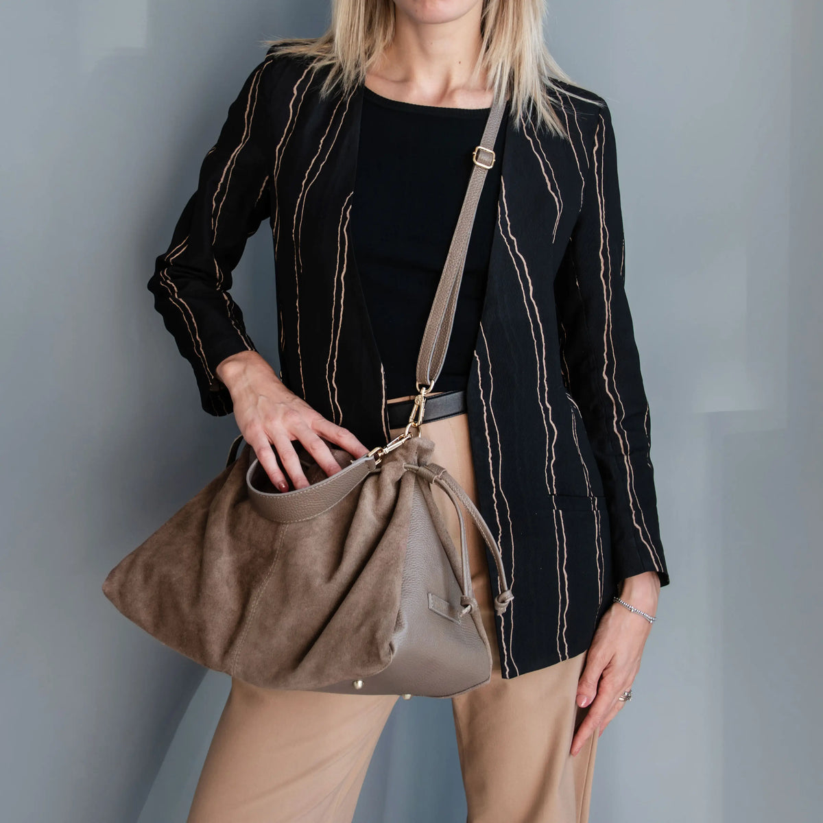 A woman wearing a black striped blazer, black top, and beige trousers carries a large taupe-brown suede shoulder bag with gold-tone hardware against a gray wall.