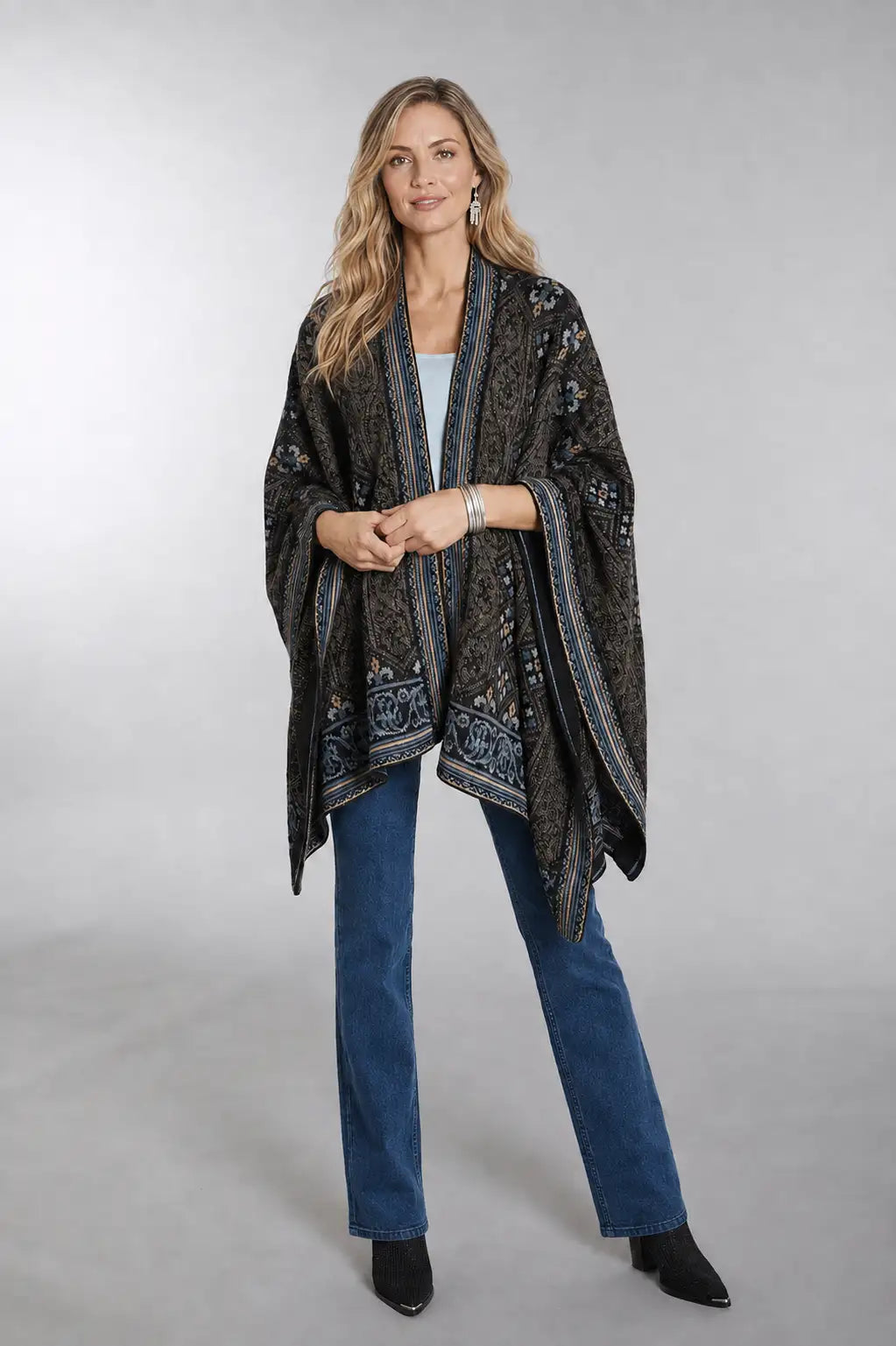 A woman modeling a dark patterned shawl wrap with blue and beige borders, worn over a white top and blue jeans against a light gray studio background.