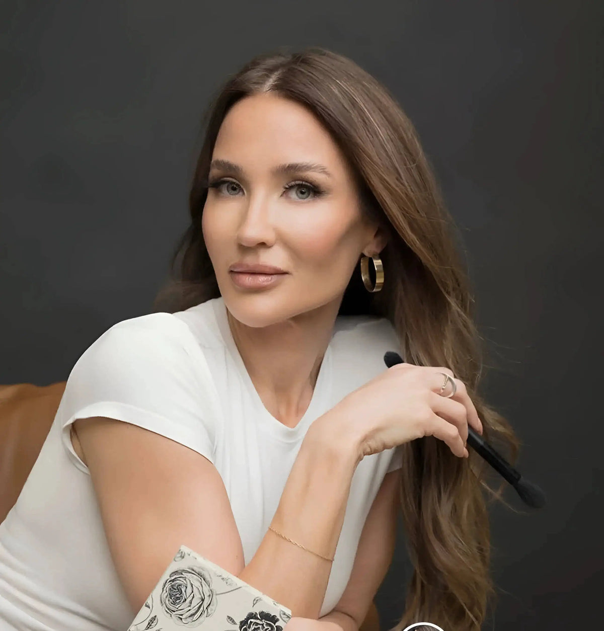 A woman with long brown hair, wearing a white t-shirt and gold hoop earrings, holding a makeup brush against a dark gray background.