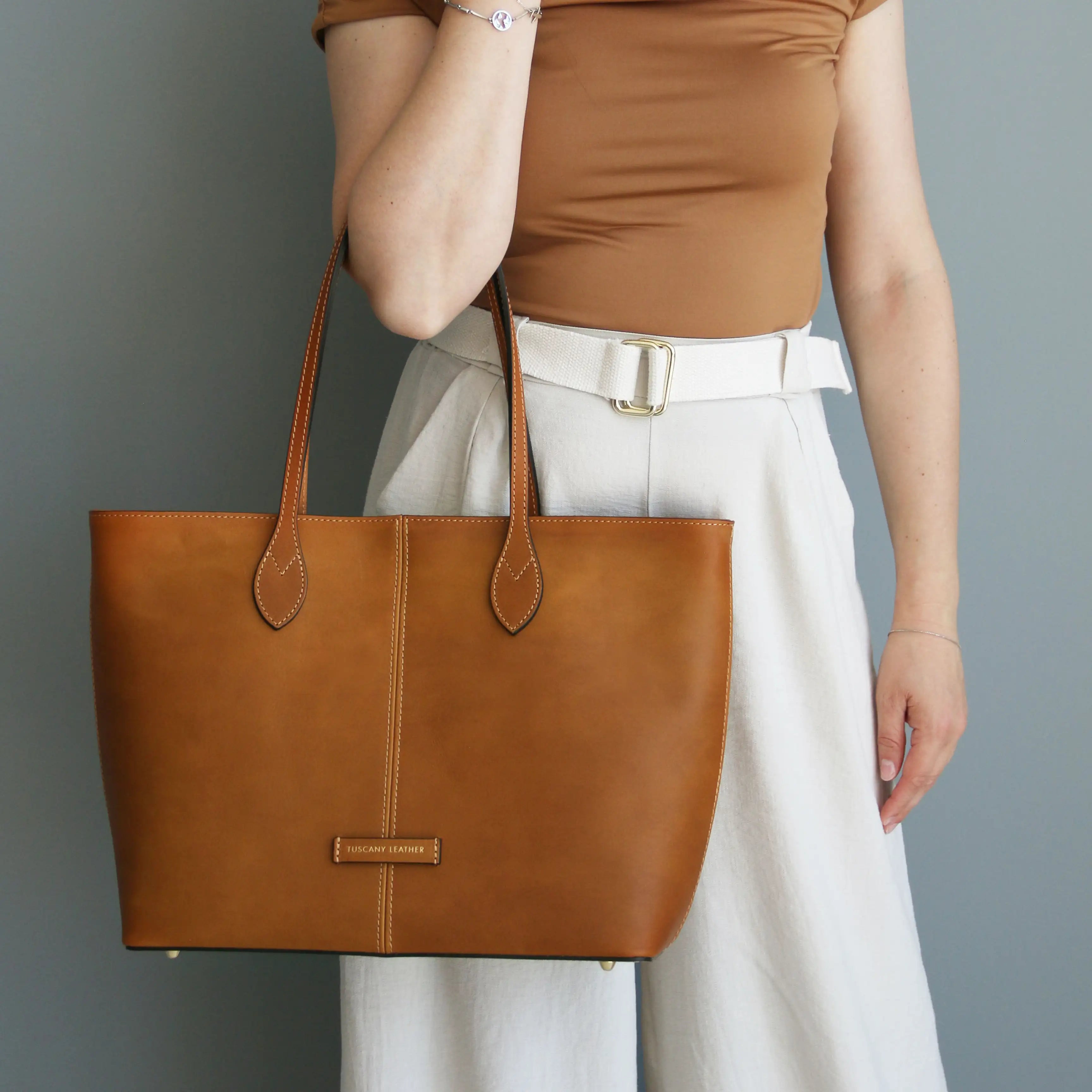 A woman is carrying the Natural Essenza Shoulder Bag wearing white pants and a cognac colored shirt