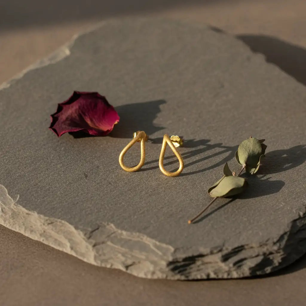 A pair of gold teardrop-shaped drop earrings displayed on a textured gray stone slab with dried rose petals and a small green bud under warm sunlight.