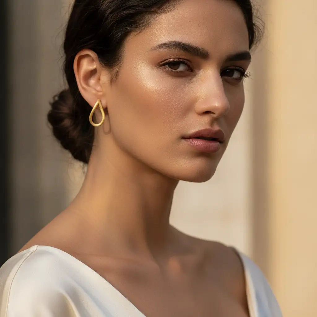 A close-up portrait of a woman wearing a delicate gold teardrop hoop earring, with her hair in a low bun and a white top against a softly blurred beige background.