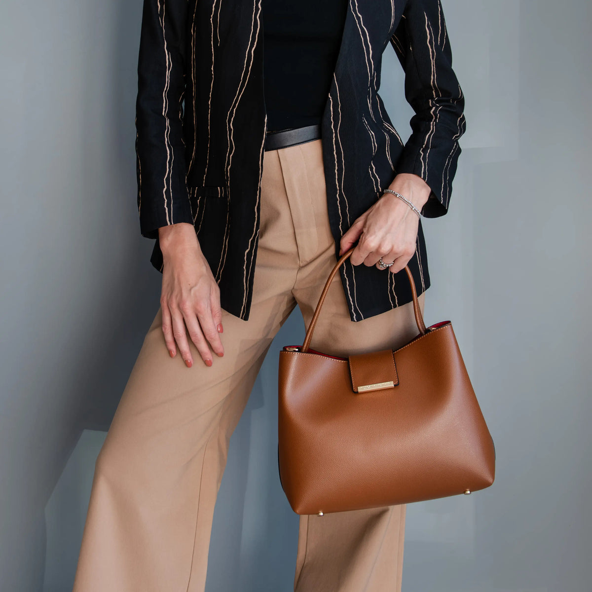 A person wearing a black striped blazer, black top, and beige wide-leg trousers holds a structured cognac leather Clio handbag with gold-tone hardware against a gray studio background.