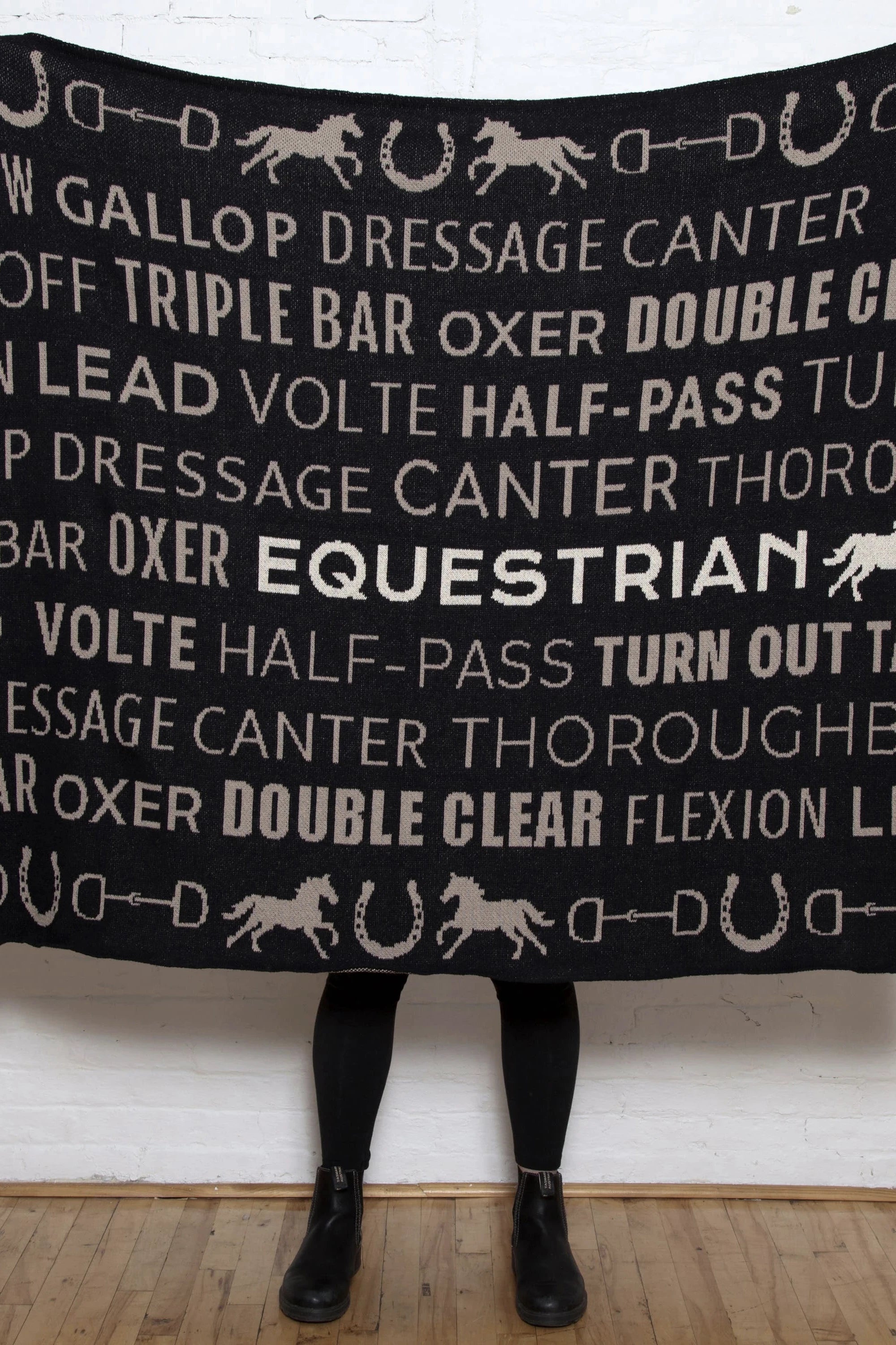 A woman is holding the Equestrian Eco Text Throw fully opened in front of her and all you see are her black boots.