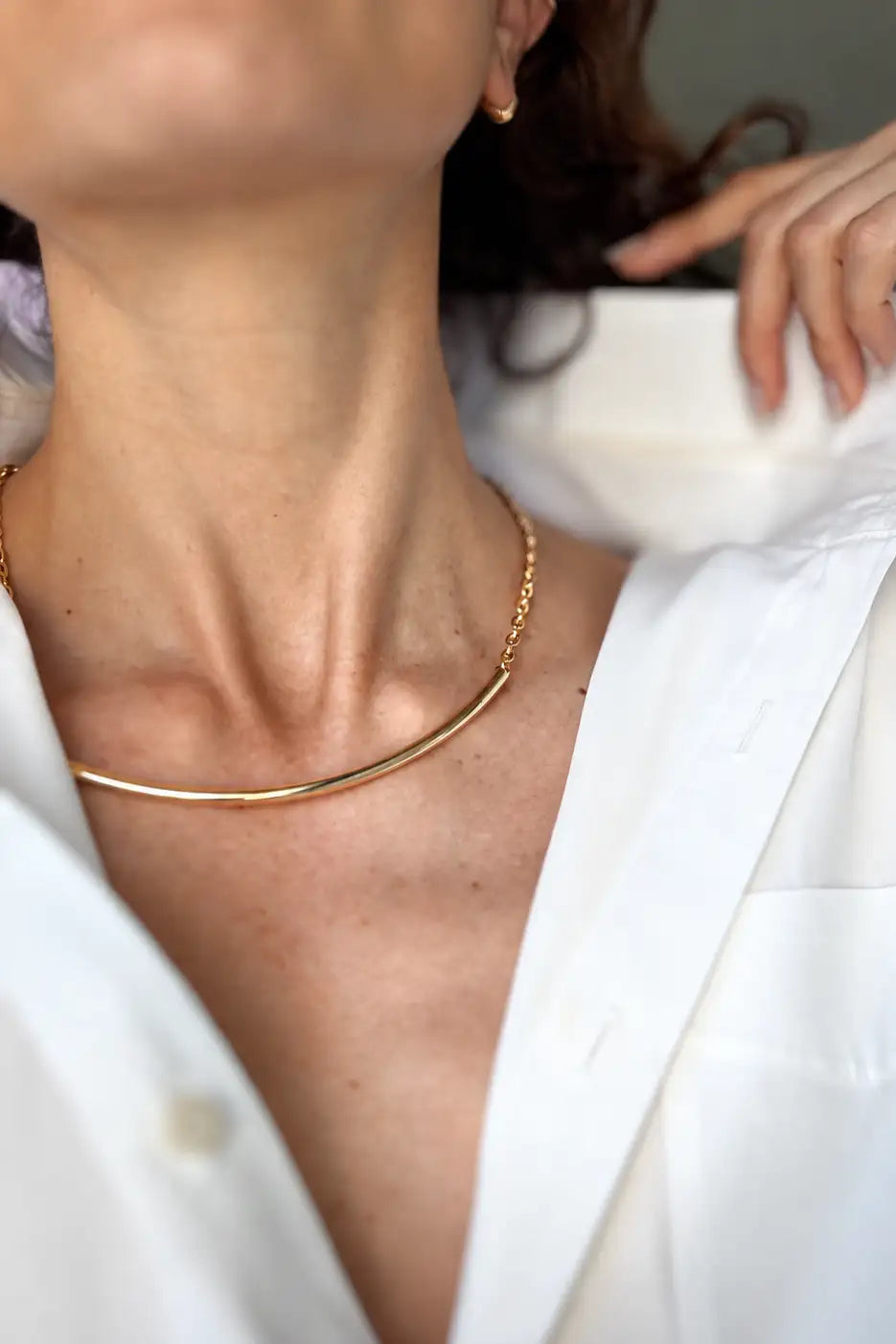 A close-up lifestyle shot of a model wearing a delicate, curved gold necklace over a white collared shirt.