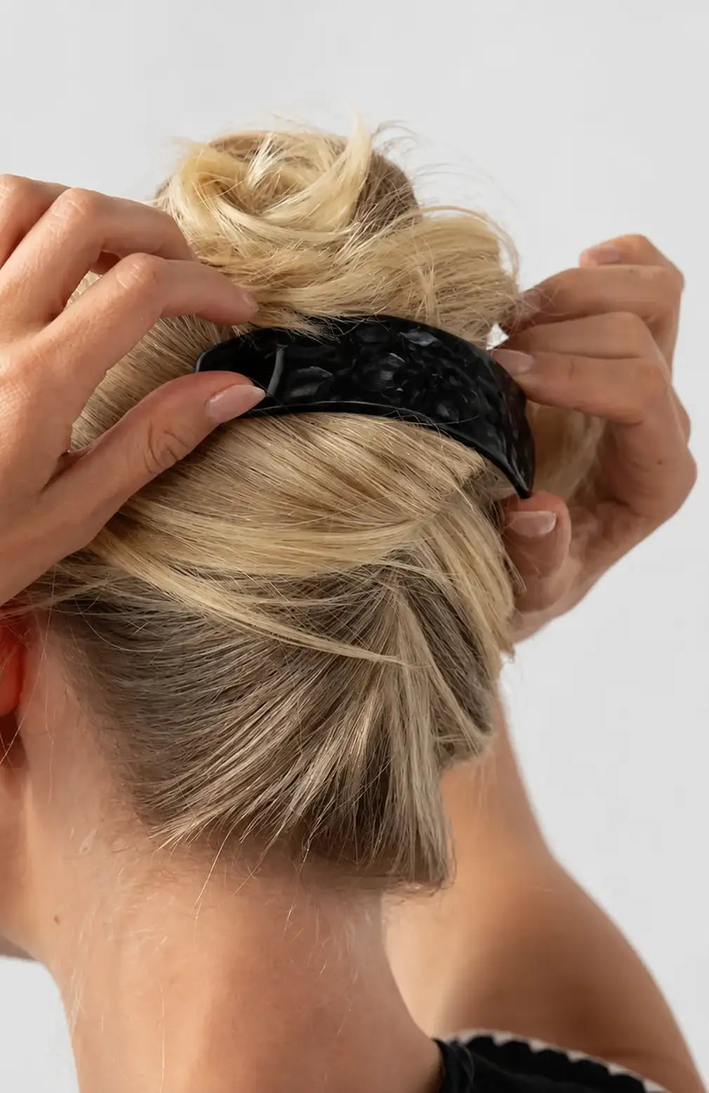 A close-up of a woman from the back, adjusting a black textured hair claw clip into a blonde bun.
