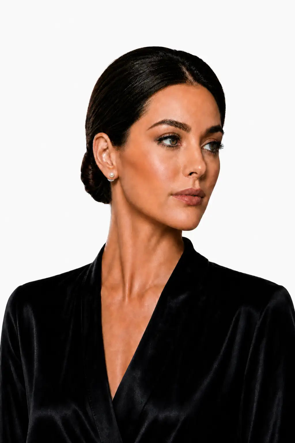 A woman with dark hair styled in a low bun wears a black satin-like wrap top and a small pearl stud earring, photographed against a plain light gray background.