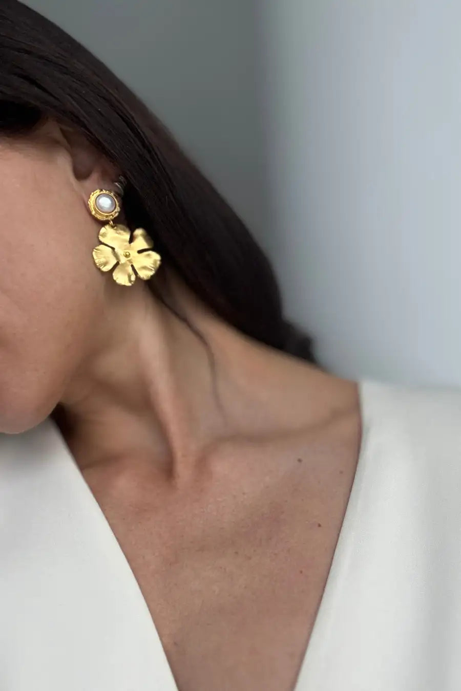 A close-up of a model wearing a gold four-leaf clover dangle earring with a white pearl at the top, paired with a white V-neck top.