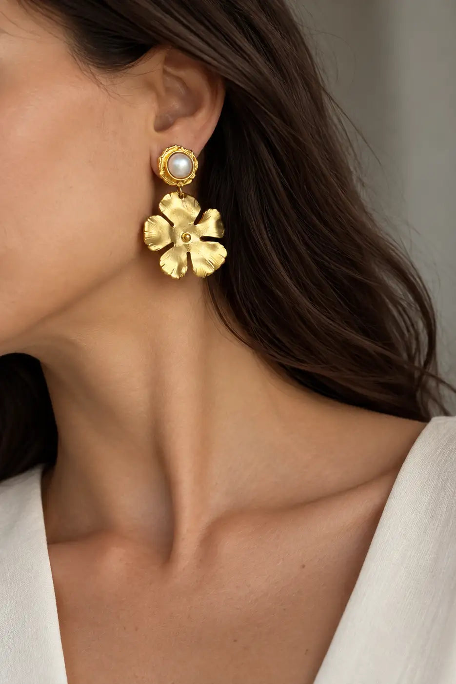 A close-up of a model wearing a gold flower-shaped drop earring with a white pearl at the top, paired with a white top.