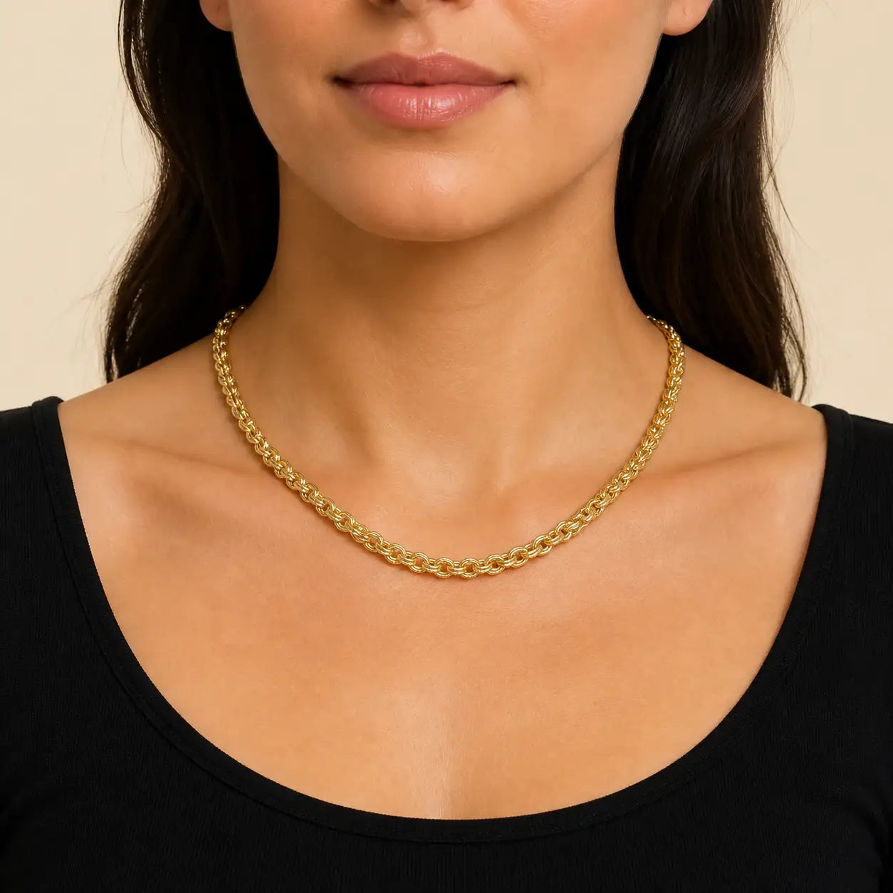 A close-up of a model wearing a gold chain necklace with a thick, textured curb link design, paired with a black scoop-neck top against a warm beige background.