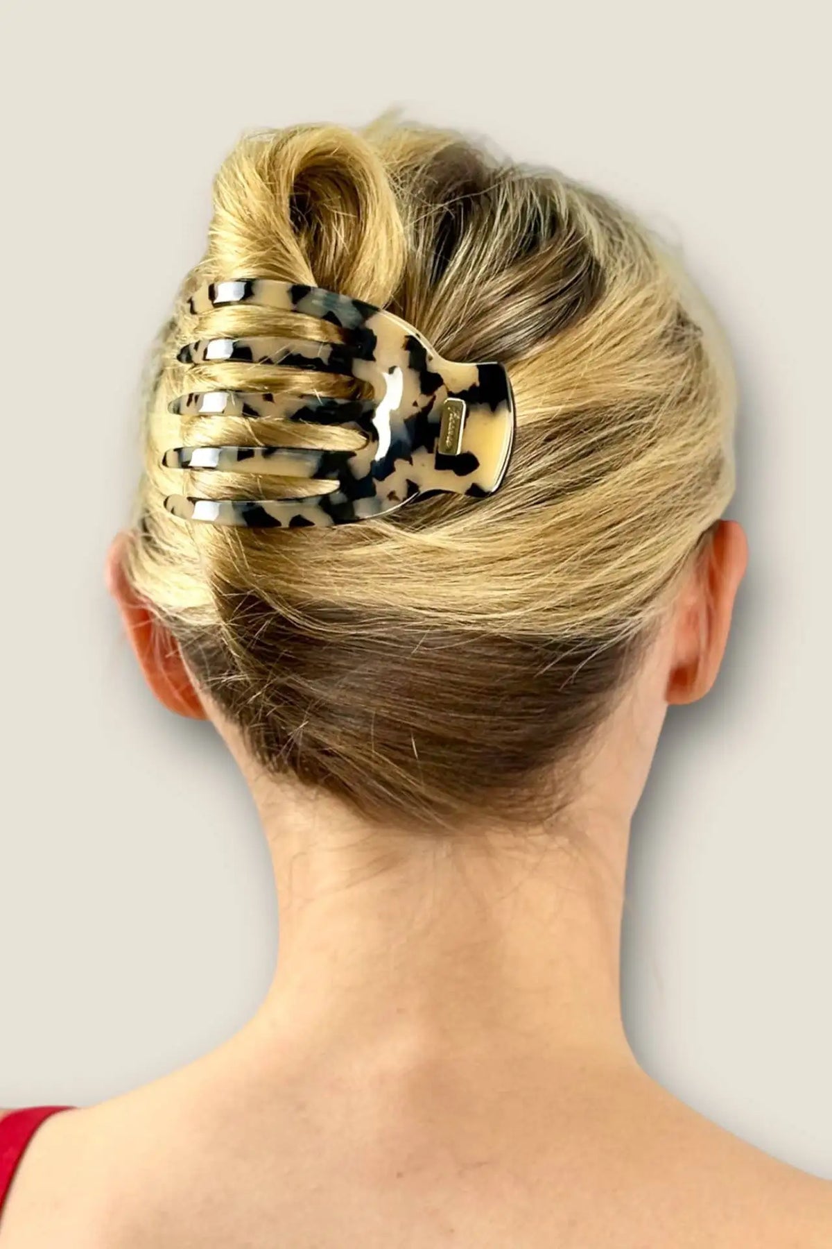 A blonde woman shown from the back wearing a tortoiseshell hair claw clip holding her hair in a low bun, against a plain light gray background.