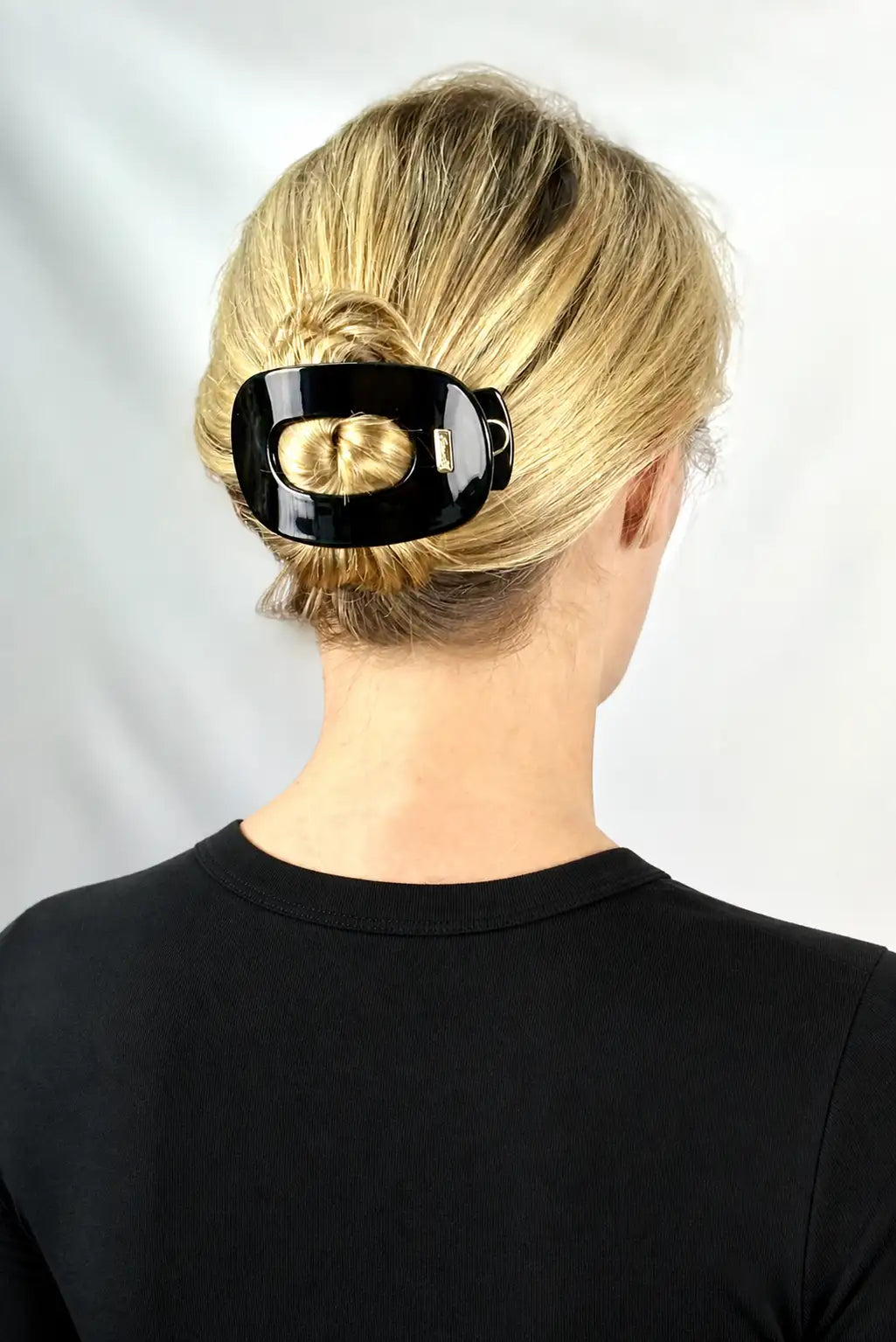 A blonde woman viewed from the back wearing a glossy black hair claw clip holding her hair in a low bun, against a light gray background.