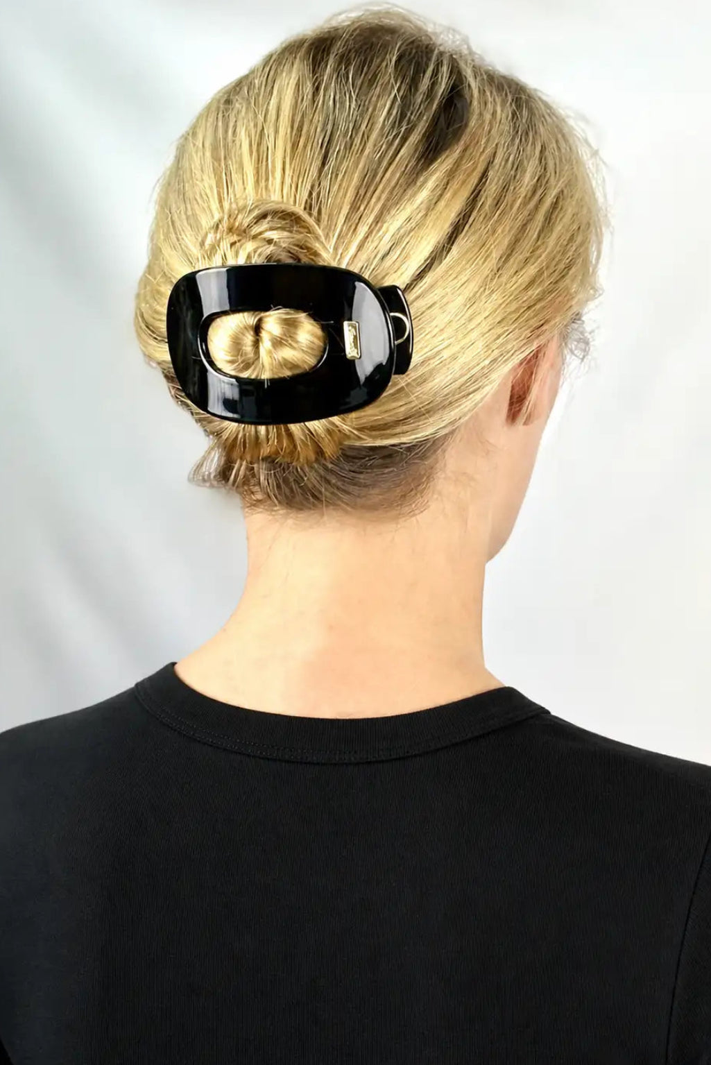 A blonde woman viewed from the back wearing a glossy black hair claw clip holding her hair in a low bun, against a light gray background.