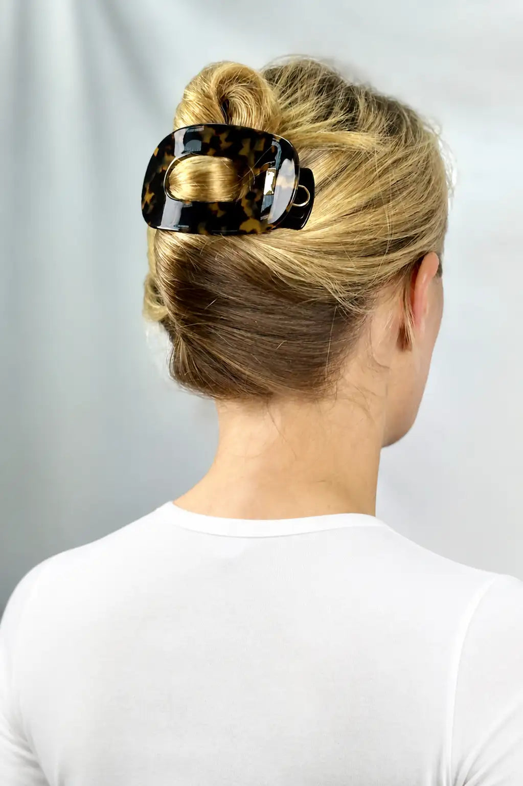 A blonde woman viewed from the back wearing a glossy black and brown tortoiseshell hair claw clip to hold her hair in a bun, against a light gray background.