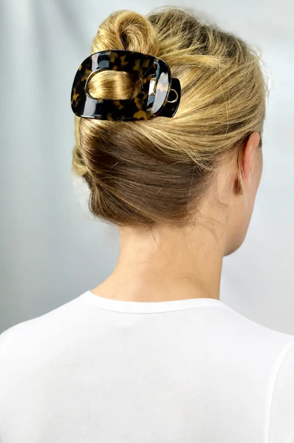 A blonde woman viewed from the back wearing a glossy black and brown tortoiseshell hair claw clip to hold her hair in a bun, against a light gray background.