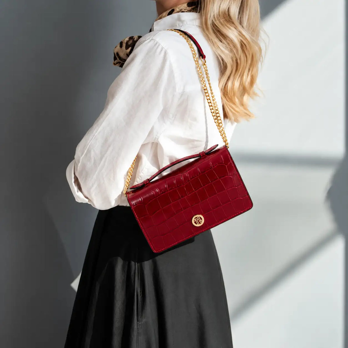 A woman wearing a white blouse and black skirt carries a structured red crocodile-embossed shoulder bag with a gold chain strap and a gold circular logo clasp, photographed against a gray wall with strong sunlight and shadows.