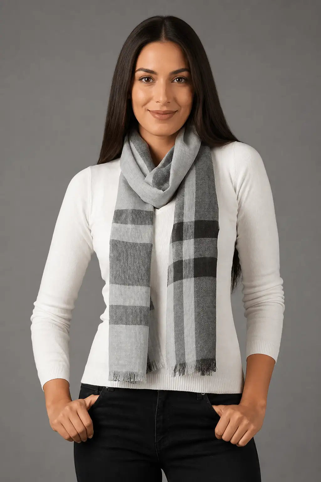 A woman modeling a gray and white plaid scarf with fringed edges, worn over a white long-sleeve top and black pants against a solid gray studio background.
