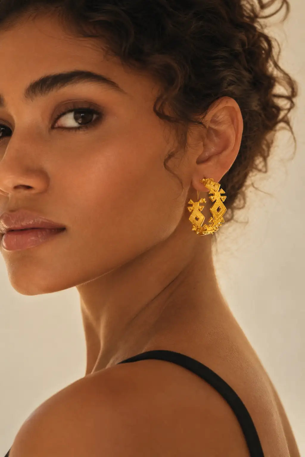 A close-up of a woman wearing a pair of gold hoop earrings with a geometric, textured pattern, set against a warm beige background.