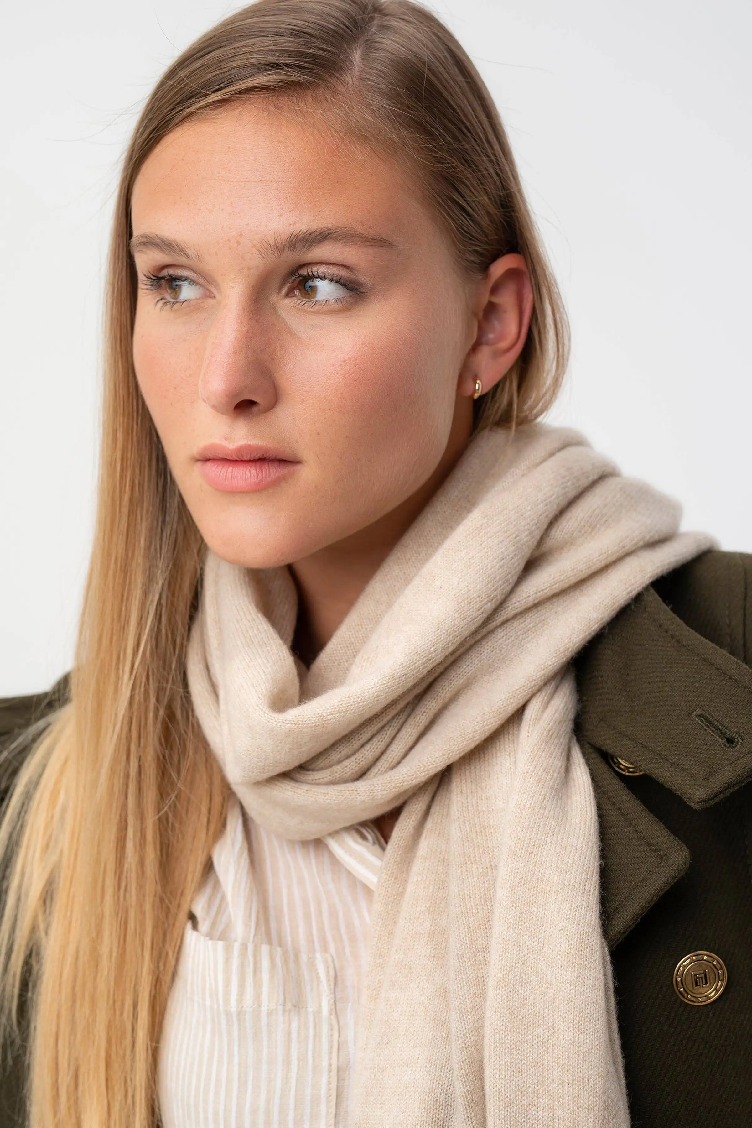 A close-up portrait of a woman wearing a soft beige-colored knit scarf wrapped around her neck, paired with a white ribbed top and an olive green jacket. She is looking off-camera against a clean, light off-white background.