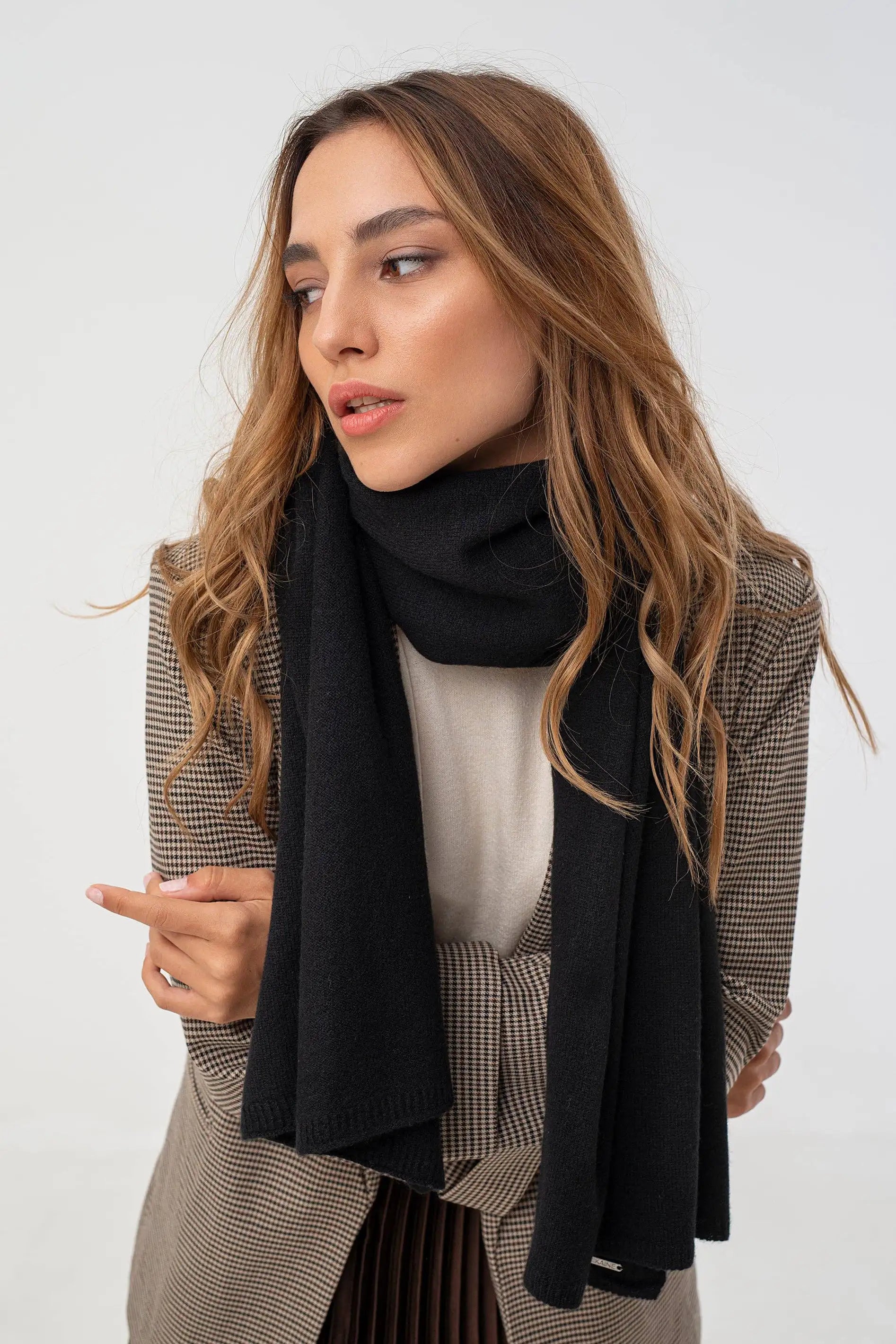 A woman with long wavy brown hair is shown from the chest up wearing a black knit scarf, a beige and brown checkered blazer, and a cream top against a plain light gray studio background.