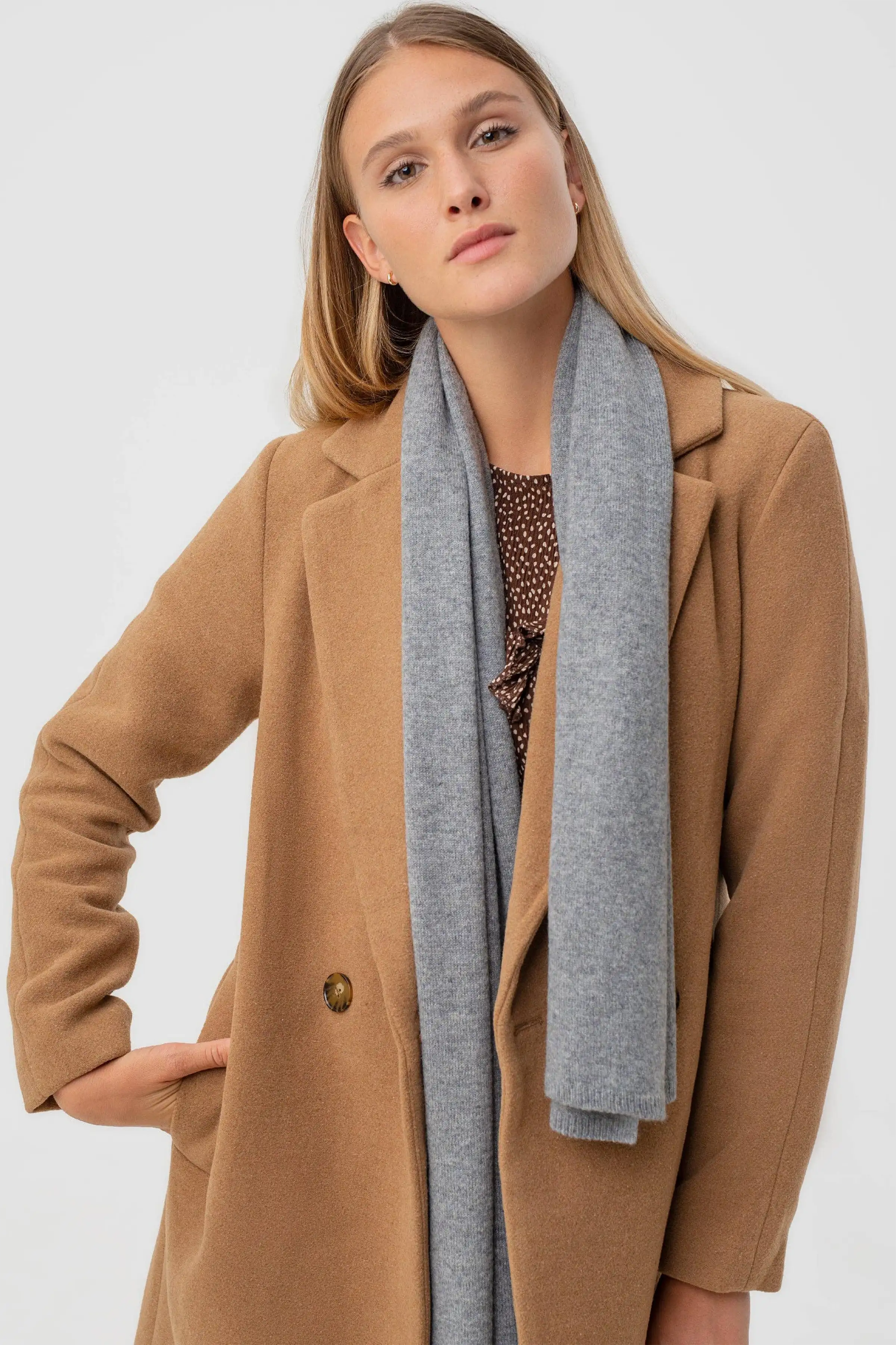 A woman wearing a camel-colored wool coat and a light gray knit scarf, posing against a solid light gray background.