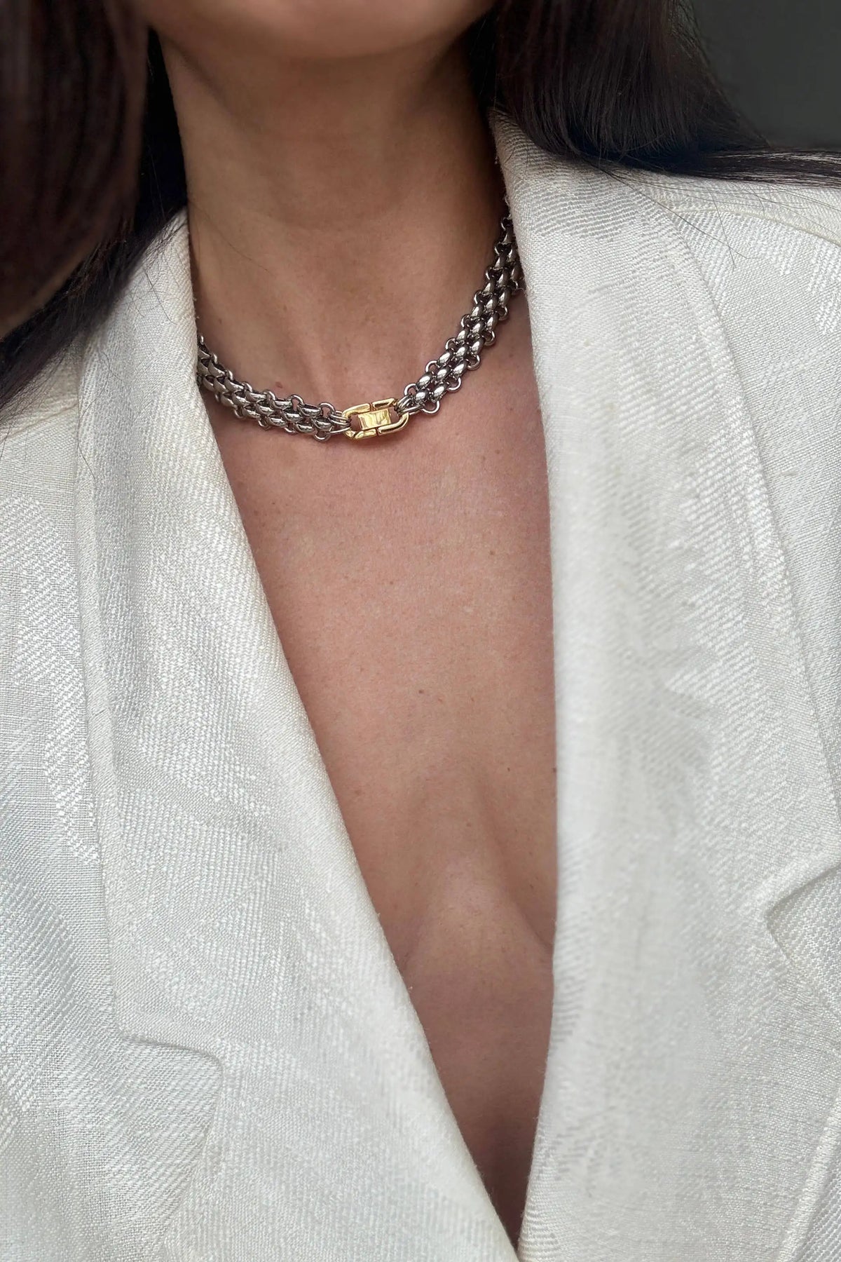 A close-up of a model wearing a silver chain choker necklace with a gold oval centerpiece, styled with a white textured blazer.