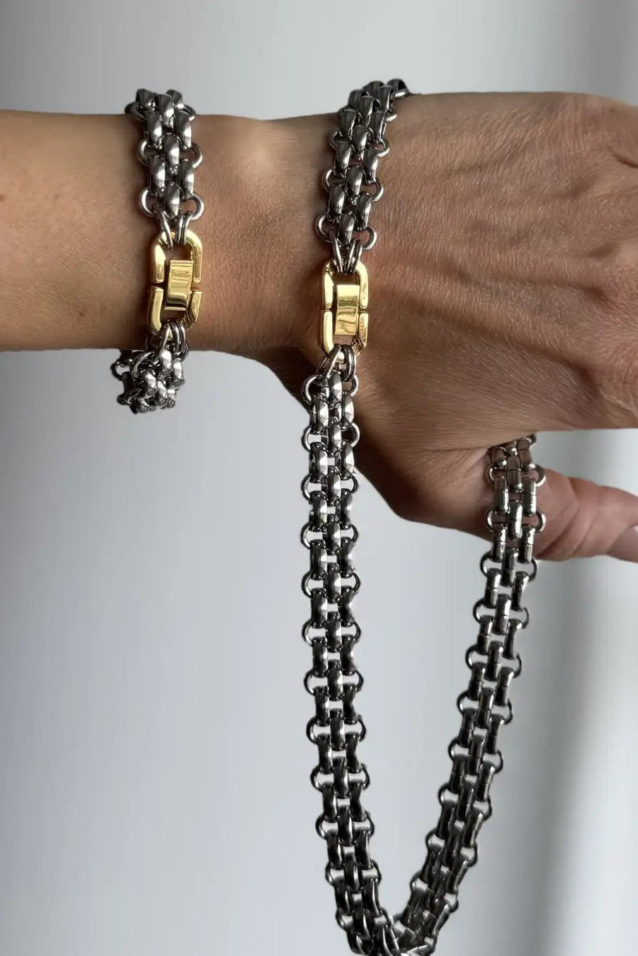 A close-up of a person's wrist and hand wearing a double-strand silver chain bracelet with gold-tone clasp connectors and a matching drop chain necklace, set against a neutral gray background.