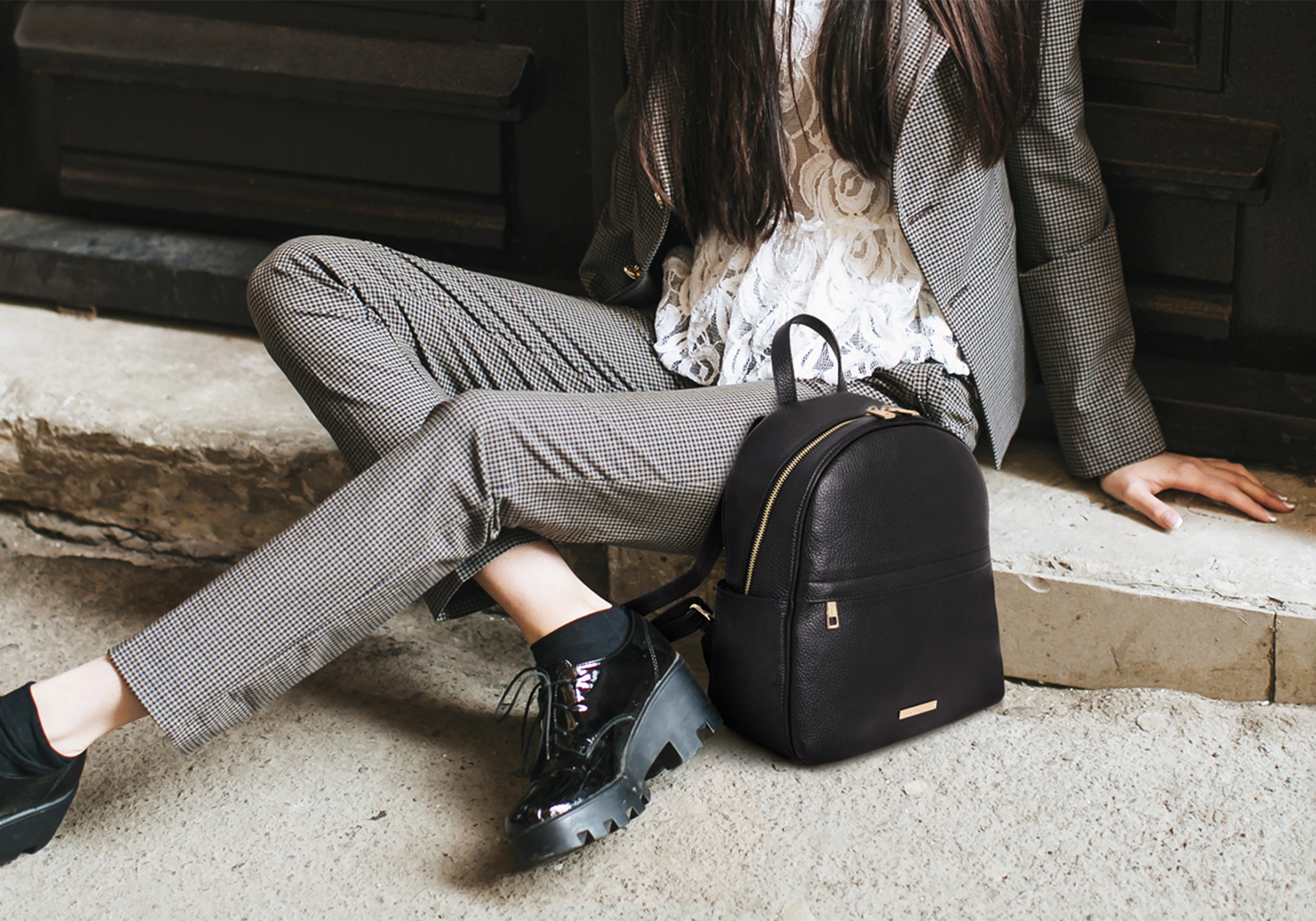 A woman sitting on a stone stoop by door with a black leather backpack purse by her left leg
