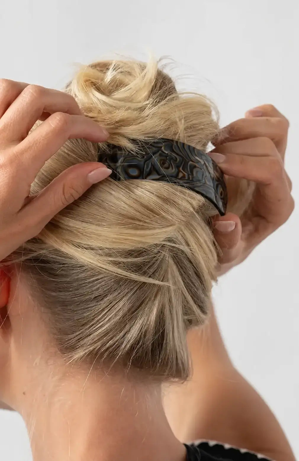 A close-up of a woman with blonde hair in a messy bun, adjusting a wide black hair barrette with a black swirl pattern against a light gray background.