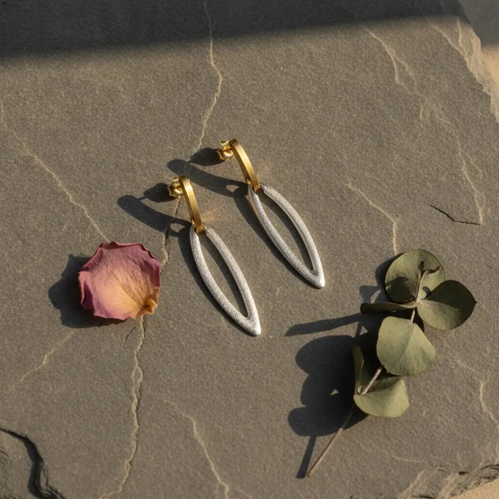 A pair of gold hoop earrings with textured silver teardrop dangles, styled on a textured stone surface with a dried rose petal and green leaves.