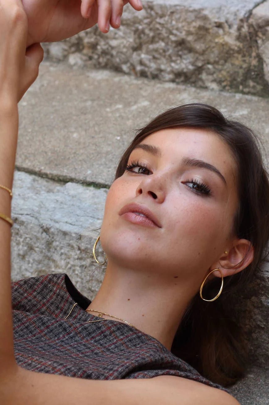 A female model is wearing the Catalonia Oval Wavy Hoop Gold Earrings. He hair is in a bun and she is laying down showing both earrings.