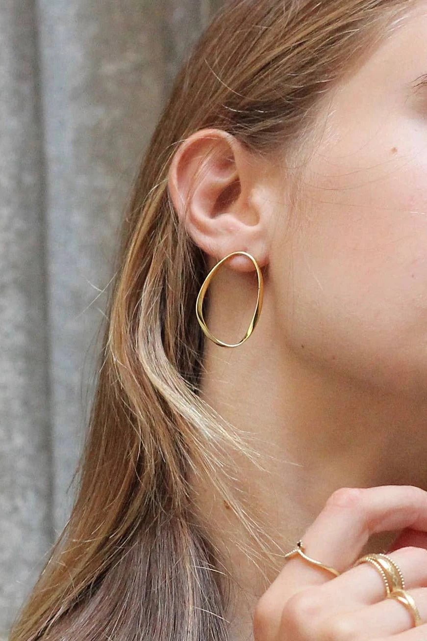 A female model is wearing the Catalonia Oval Wavy Hoop Gold Earrings. He hair is straight and she is looking to the right showing her right earring.