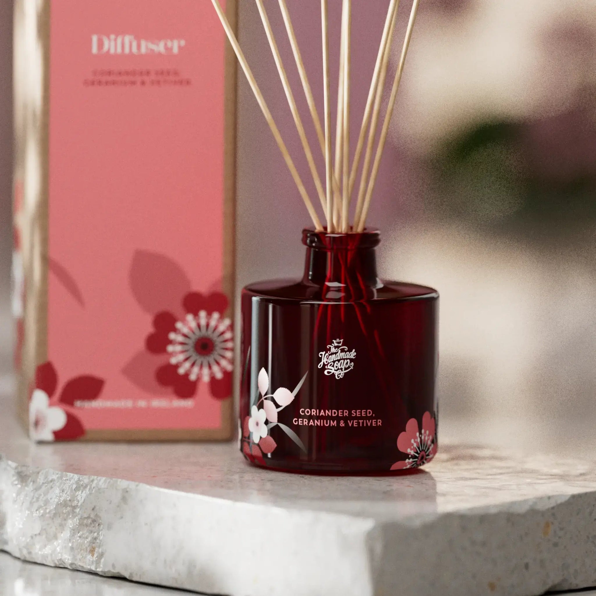 Red diffuser bottle with floral design on a marble surface, with a pink box in the background.