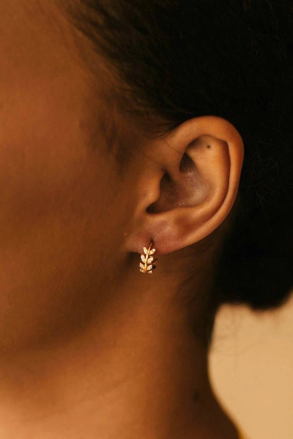 A model is wearing the Dancing Leaves Cubic Zirconia Huggie Hoop Earrings in Gold on her left ear.