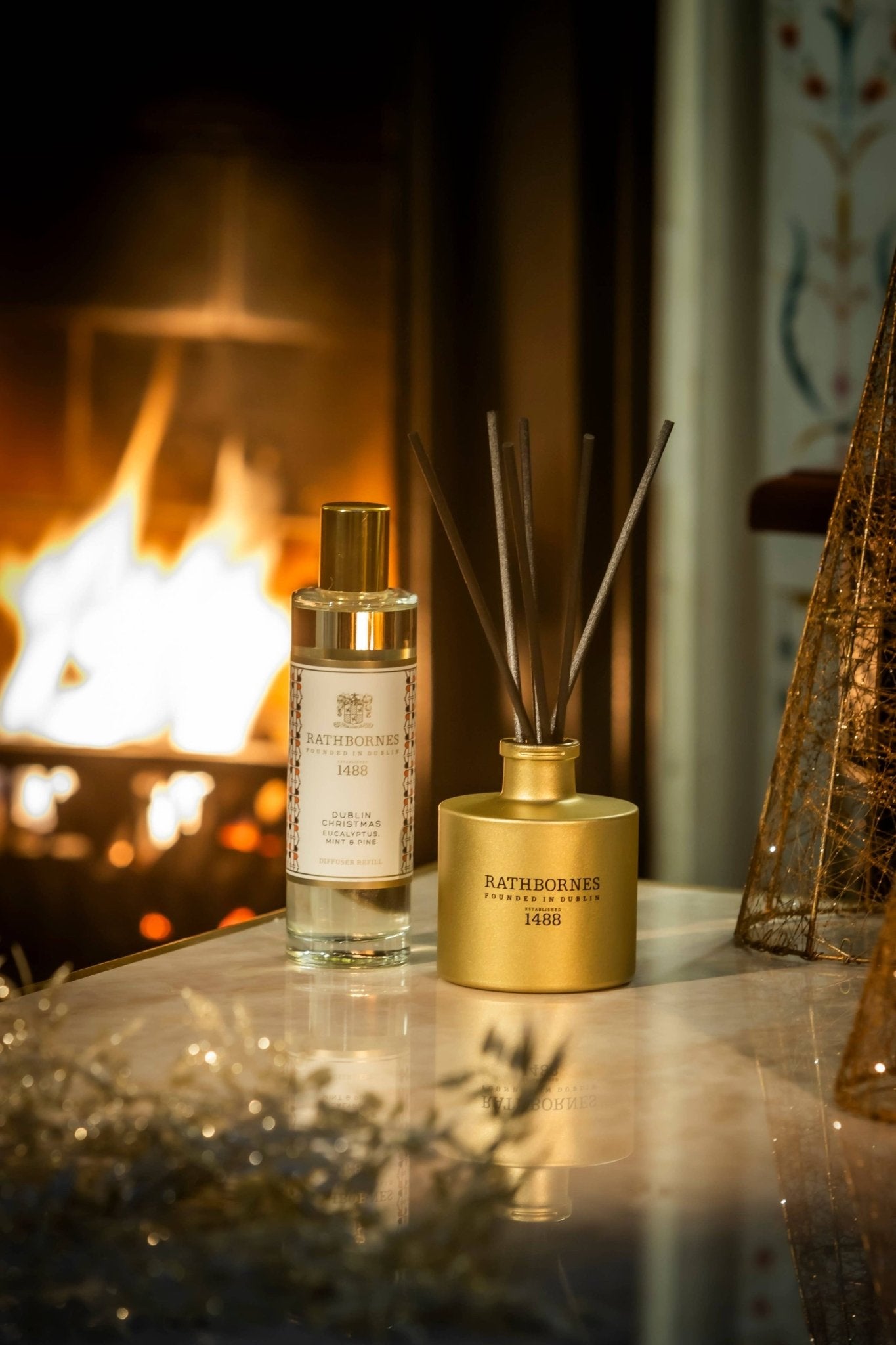 Dublin Christmas Scented Reed Diffuser (Eucalyptus, Mint & Pine) gold bottle and refiller bottle on a livingroom table near a fire