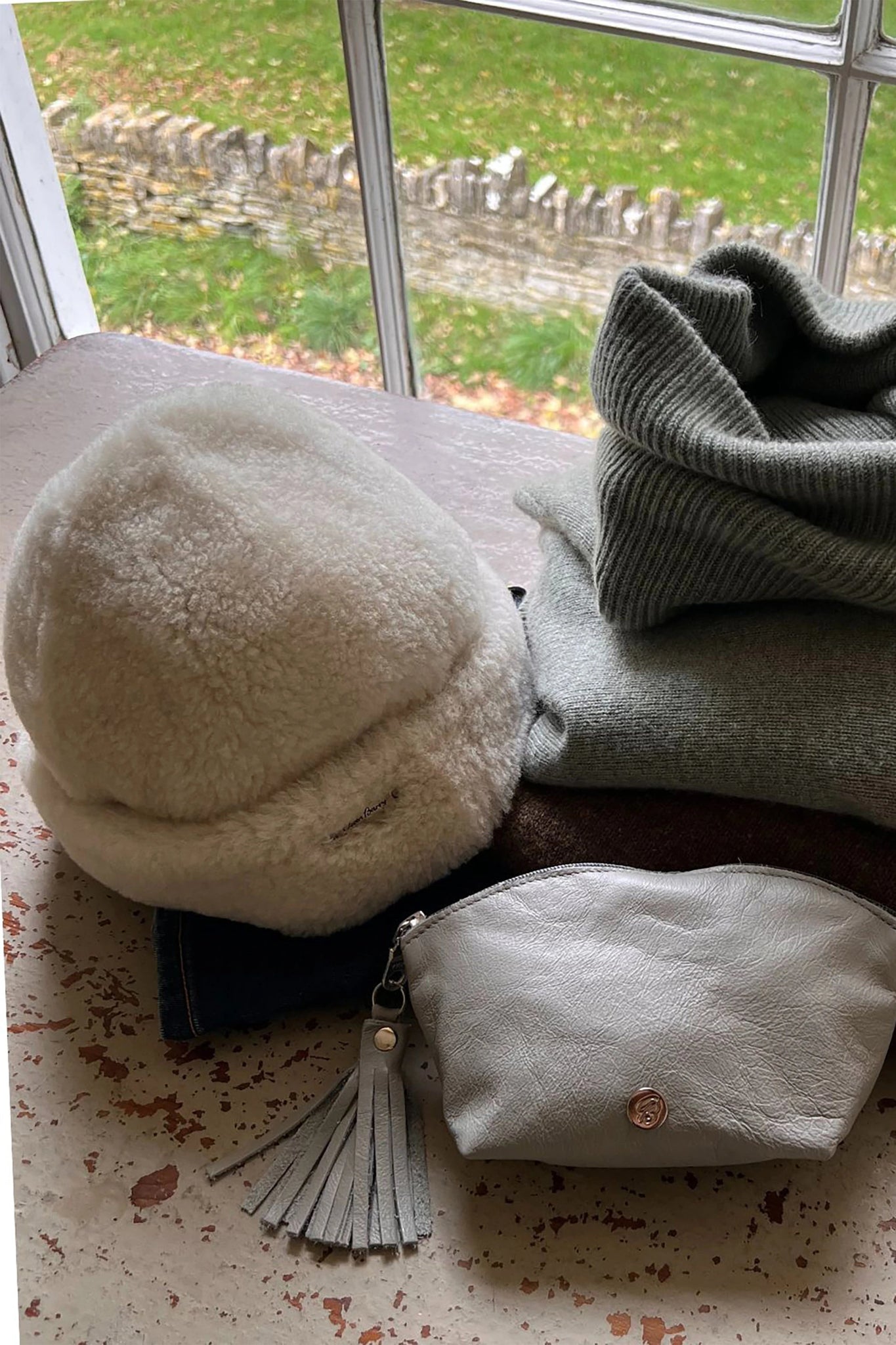 English Sheepskin Pull On Beanie in Cream on a table next to a white cosmetic bag and a gray and green sweater